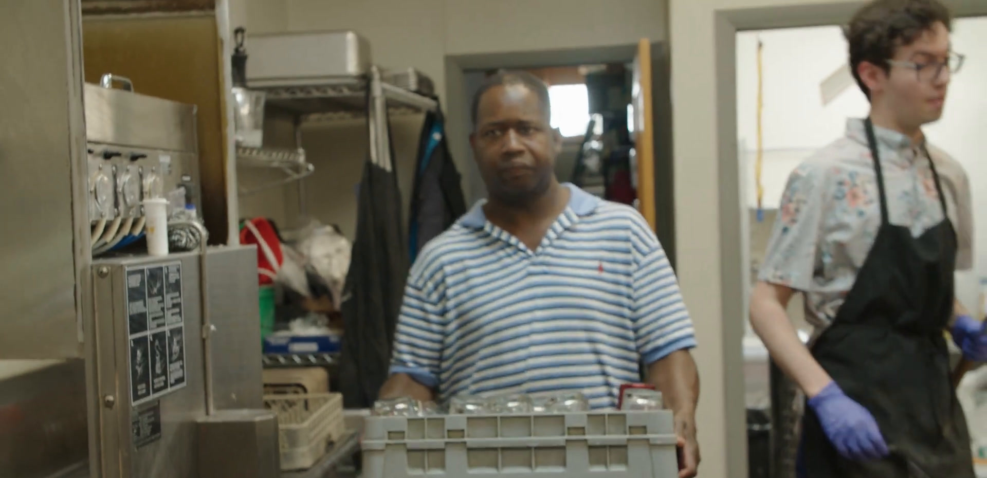 Aaron in a striped polo carrying a rack of glassware through the bar.